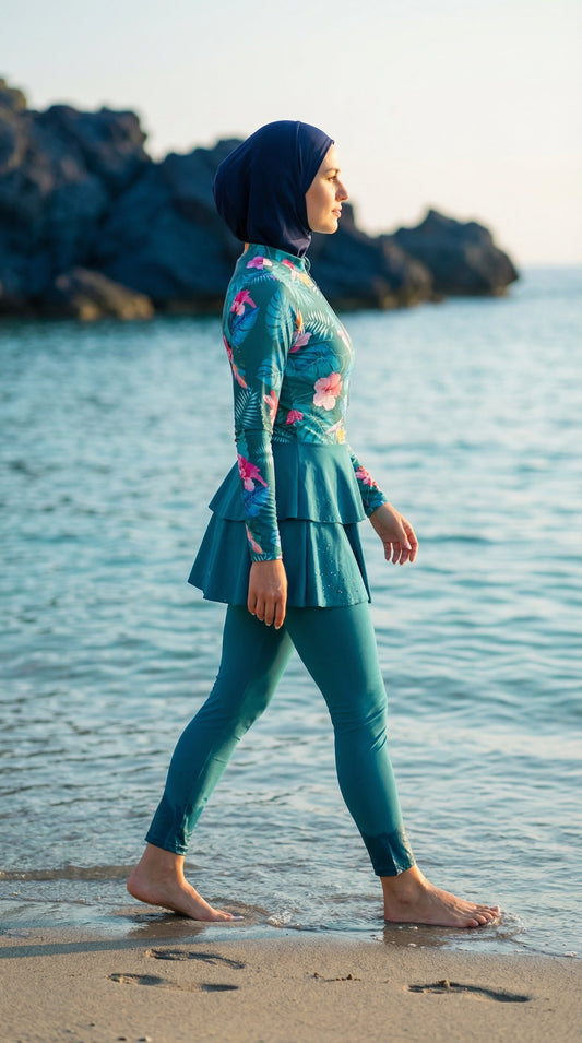 Aria Marble – Burkini Damen, Woman in a teal swimsuit with pink floral patterns walking on a beach.