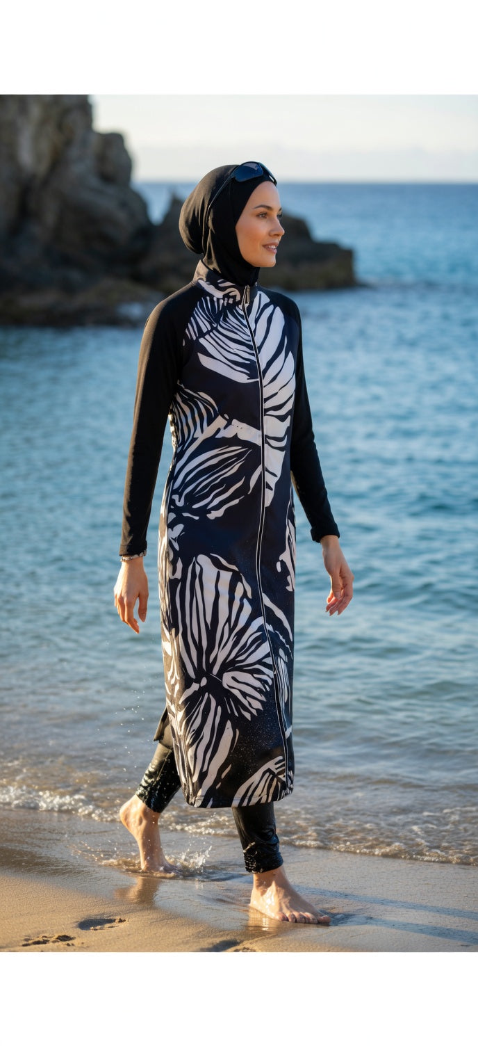 Woman in a long, dark patterned white burkini set walking on a beach with ocean and rocks in the background.