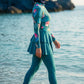 Aria Marble – Burkini Damen, Woman in a teal swimsuit with pink floral patterns walking on a beach.
