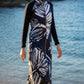 Woman in a long, dark patterned white burkini set walking on a beach with ocean and rocks in the background.